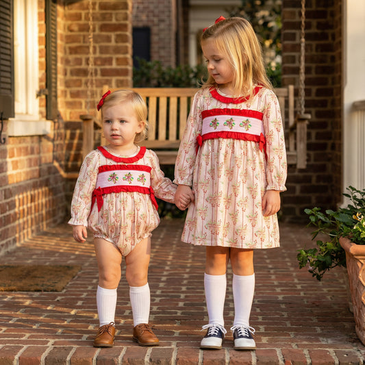 Sisters in matching Grandmillennial Holly Berry smocked dress and bubble romper holding hands on a porch.