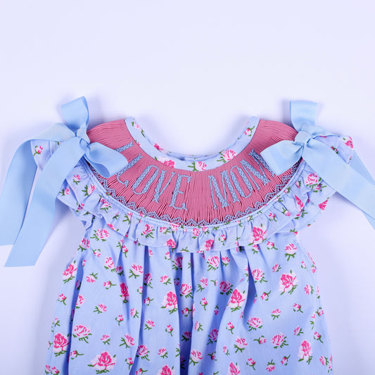 Close-up of pink hand smocked yoke with blue I Love Mom hand embroidery and shoulder bows on baby outfit.