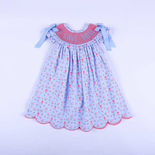 Girls light blue floral hand-smocked dress with I Love Mom embroidery, shoulder bows, and scalloped hem front view.
