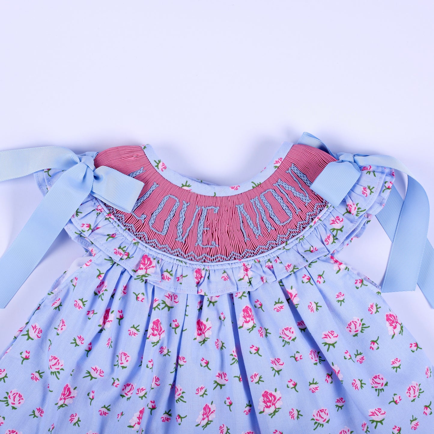 Close-up of I Love Mom hand-embroidered smocked bodice with blue ribbon bows on floral baby girl top.