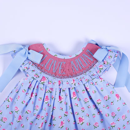 Close-up of I Love Mom hand-embroidered smocked bodice with blue ribbon bows on floral baby girl top.