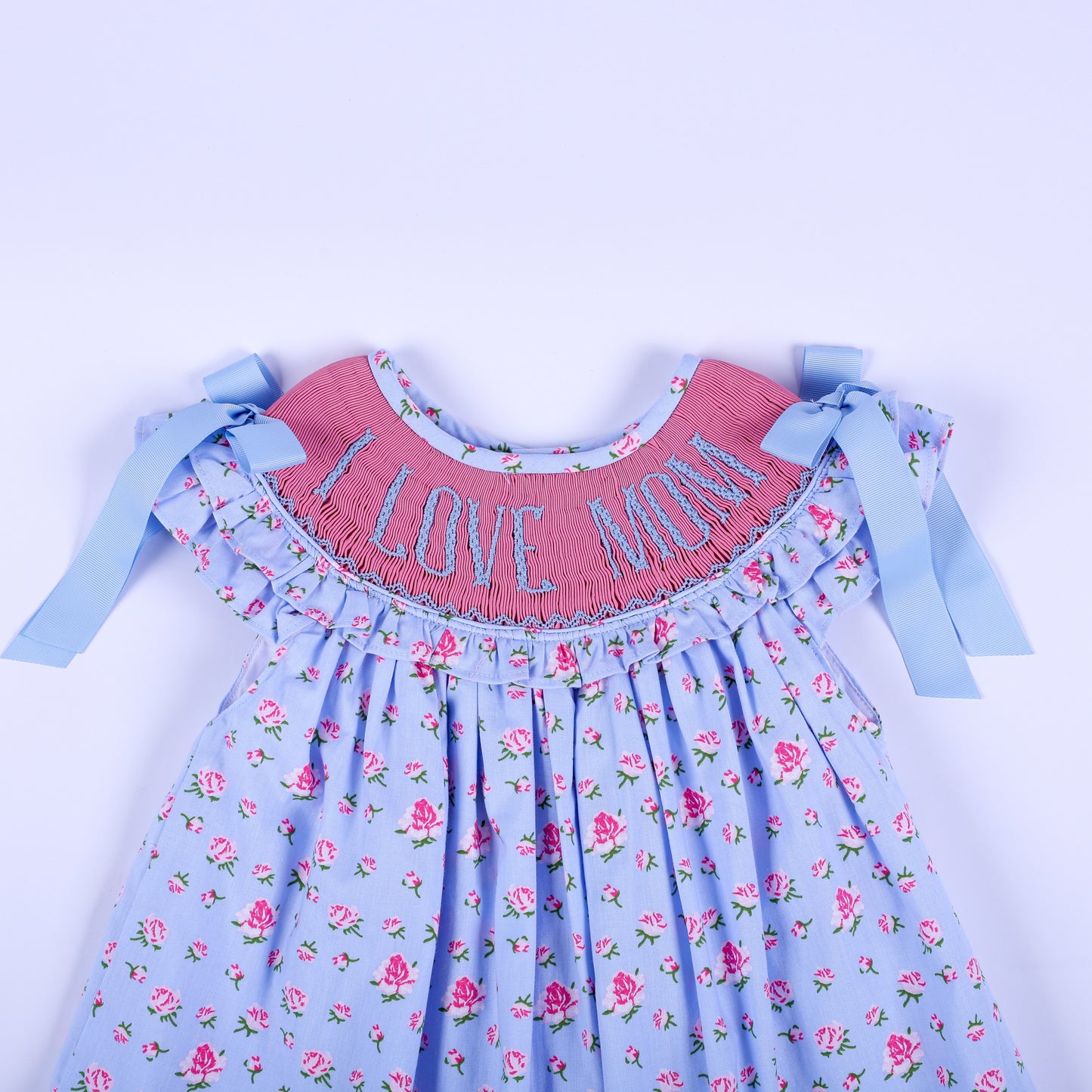 Close-up of pink hand-smocked yoke with blue I Love Mom embroidery, flutter sleeves, and shoulder ribbons on dress.