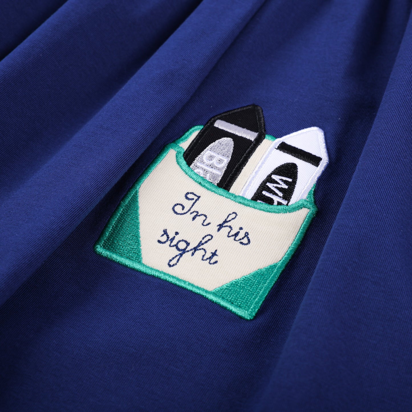 Detail of the crayon box pocket on the navy smocked dress, embroidered with 'in his sight' and holding crayons.