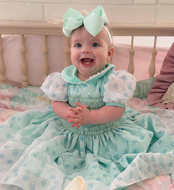 Smiling baby girl in Jenny's mint chiffon smocked floral dress with sheer puff sleeves and a matching bow headband.