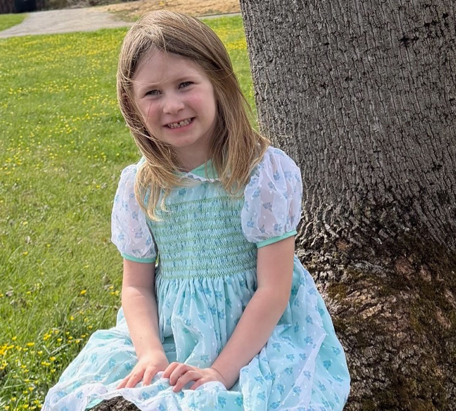 Little girl wearing Jenny's mint floral smocked dress with sheer puff sleeves sitting outside by a tree trunk.