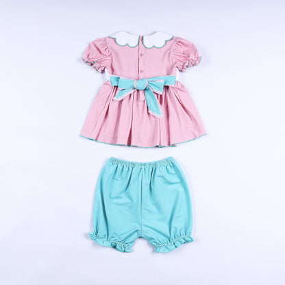 Back view of pink hand-smocked dress set showing turquoise sash bow closure and matching ruffle bloomers.