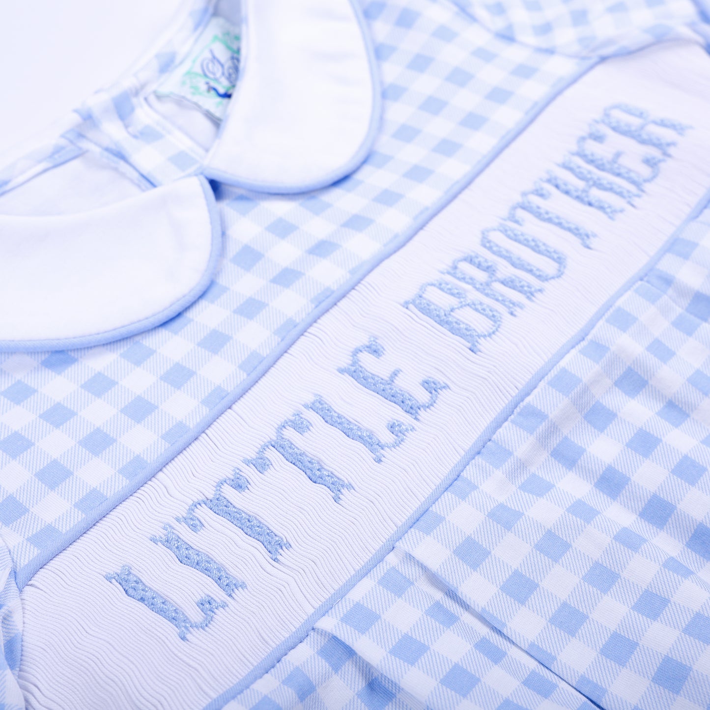 Angled close-up of hand-embroidered Little Brother text on white smocking plate of blue gingham bubble.