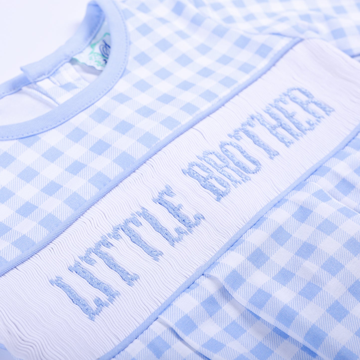 Angled close up of hand embroidered Little Brother smocked chest plate on blue gingham baby gown layette.