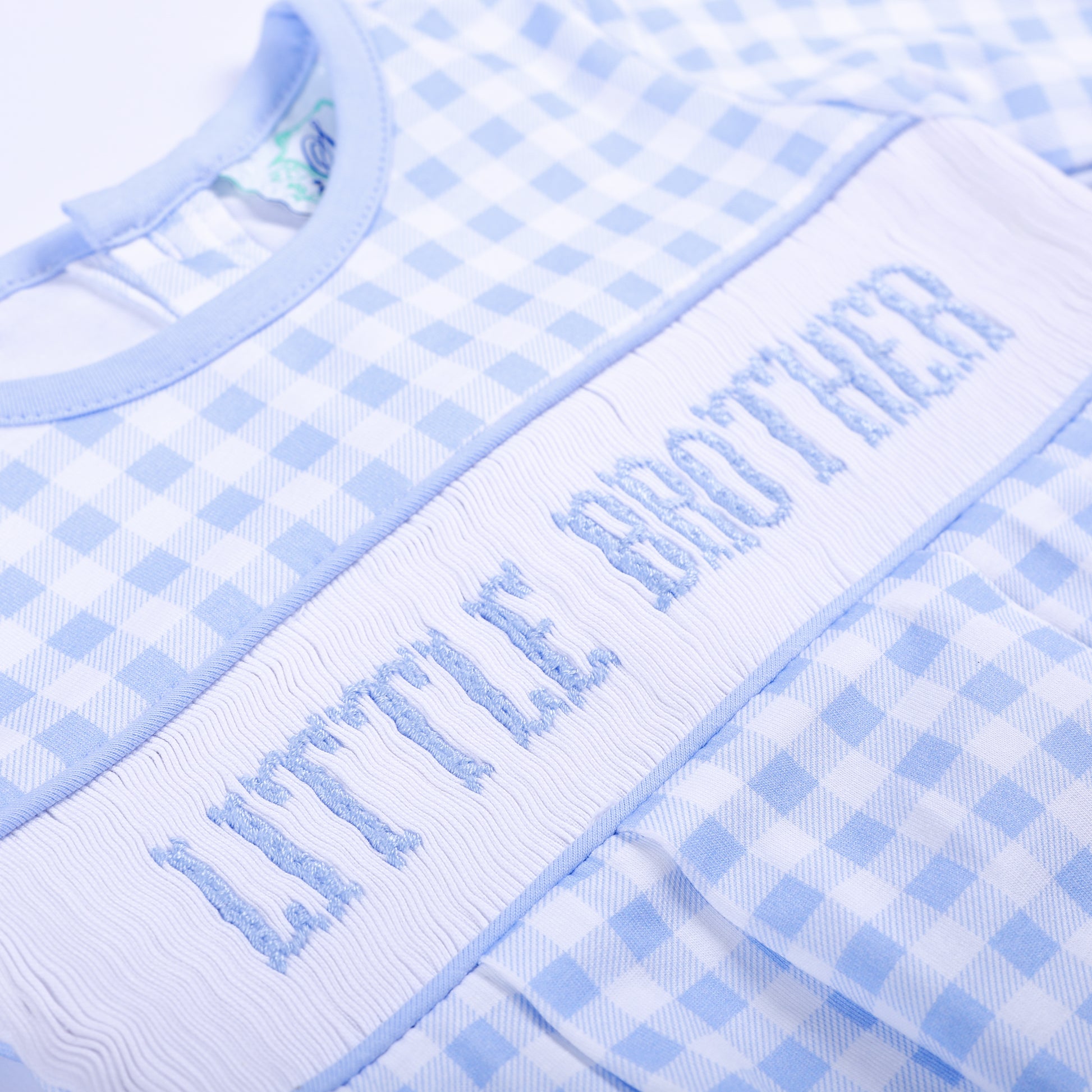 Angled close up of hand embroidered Little Brother smocked chest plate on blue gingham baby gown layette.