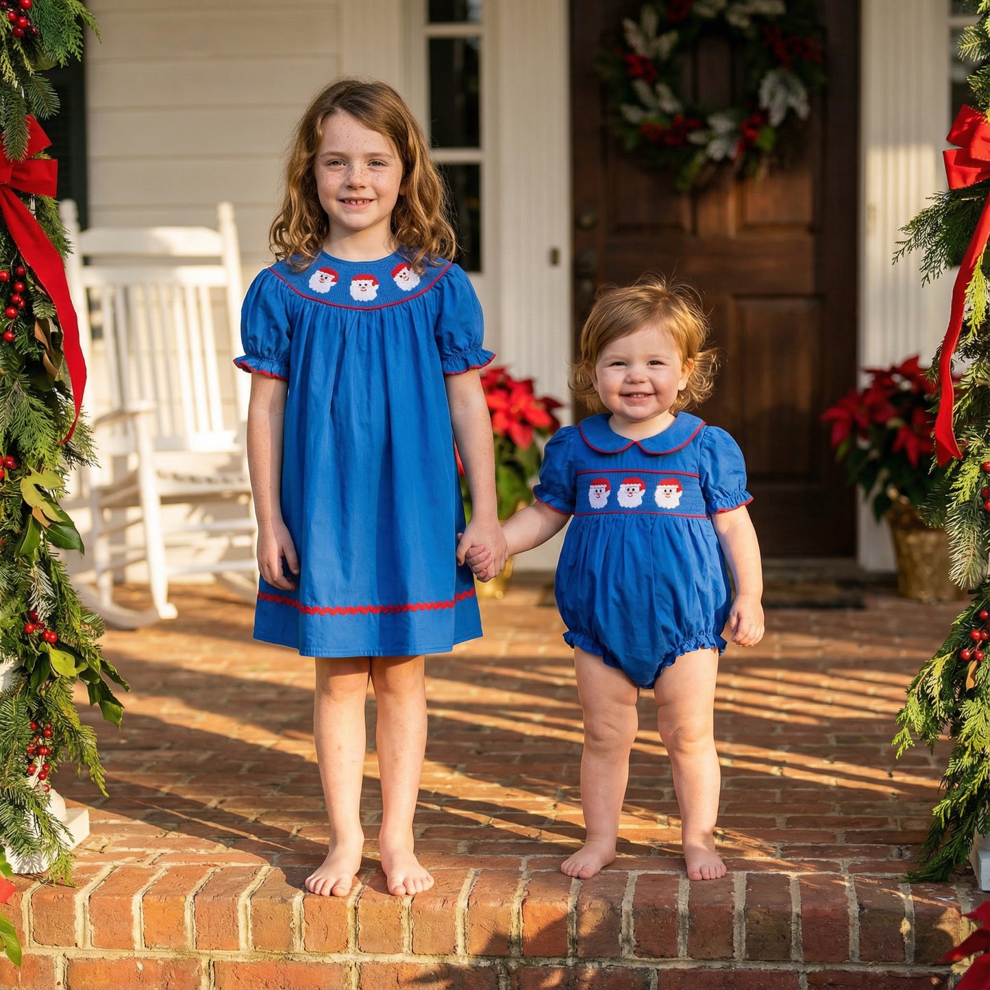 Siblings holding hands on a porch in matching navy blue smocked Santa dress and bubble romper for Christmas.