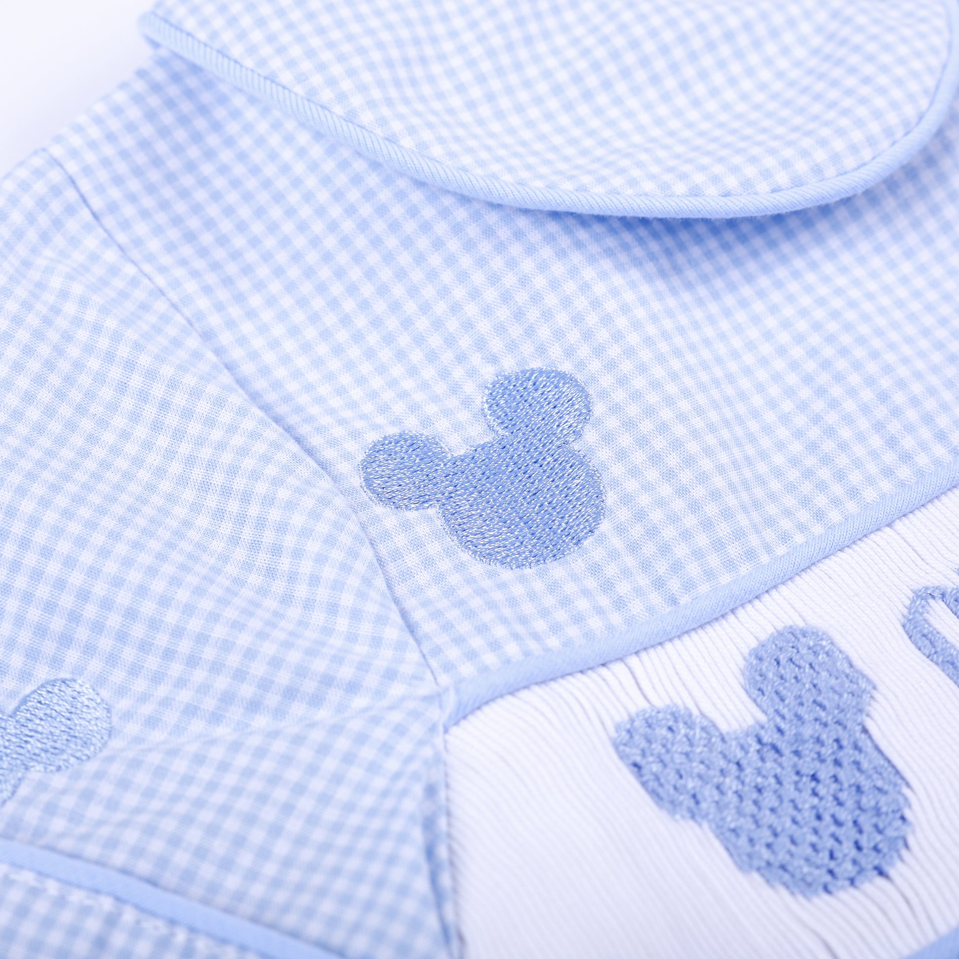 Macro view of light blue embroidered mouse motif and delicate smocked stitching on boys gingham Oh Twodles bubble.