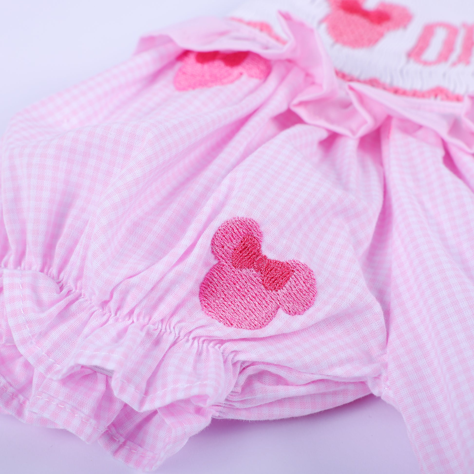 Detailed view of elastic ruffled leg opening and embroidered mouse head on pink gingham hand smocked bubble.
