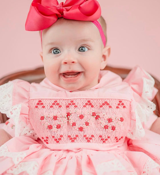 Smiling baby girl wearing a pink hand-smocked 2-piece dress with hand-embroidered details and detachable lace apron.