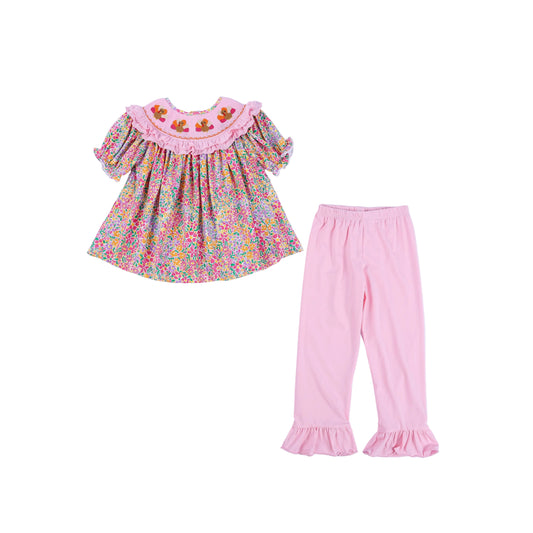Two-piece girls Thanksgiving outfit featuring a pink floral hand-smocked tunic top with embroidered turkeys and light pink ruffle-hem pants.