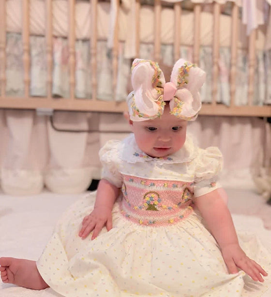 Baby girl wearing a pink flower basket hand-smocked dress with hand-embroidered details and matching bow headband.