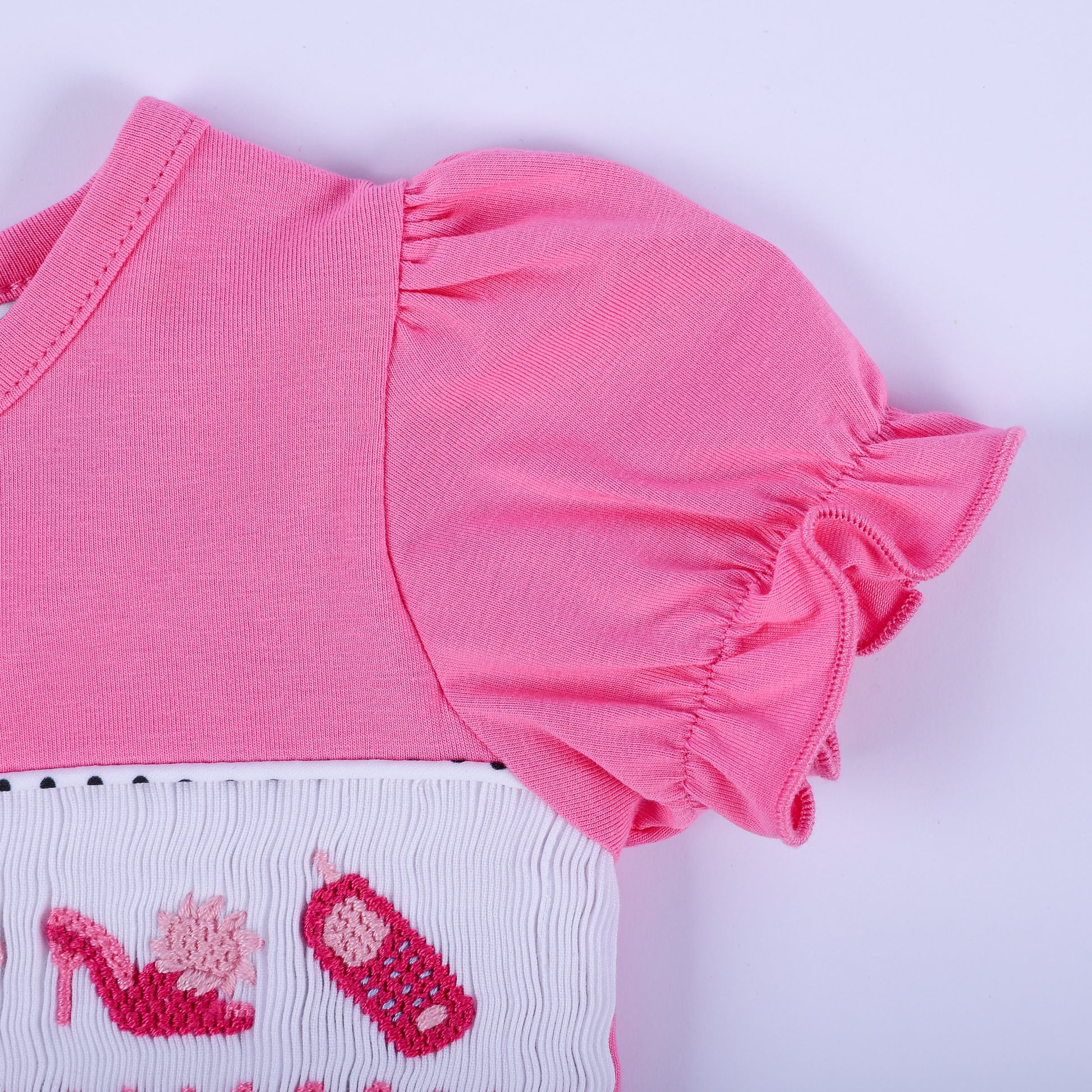 Detailed shot of pink flutter sleeve and hand-embroidered high heel with vintage cell phone on white smocked chest.