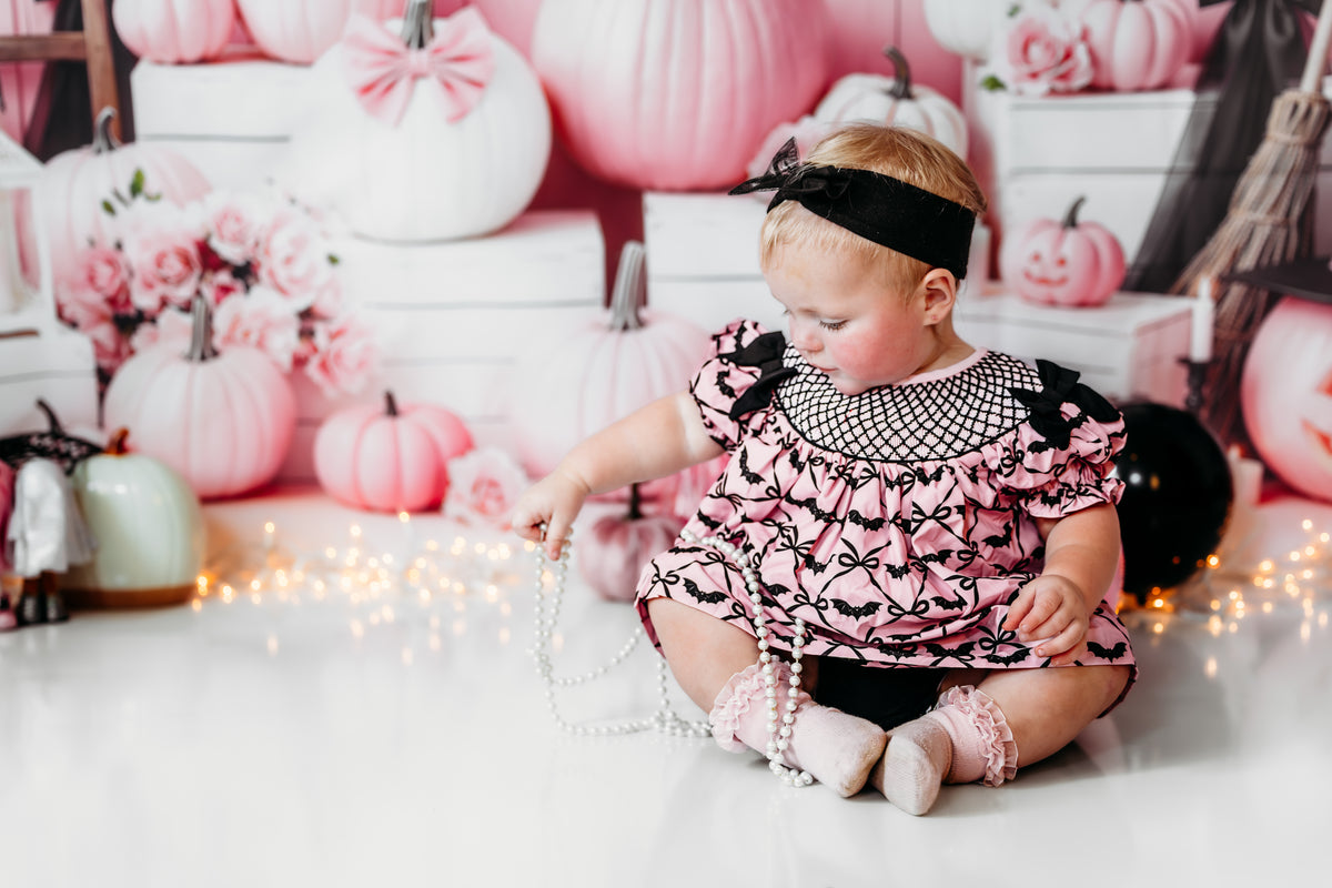 Baby girl sitting on floor in pink hand smocked bats bloomer set playing with pearls amidst fairy lights.