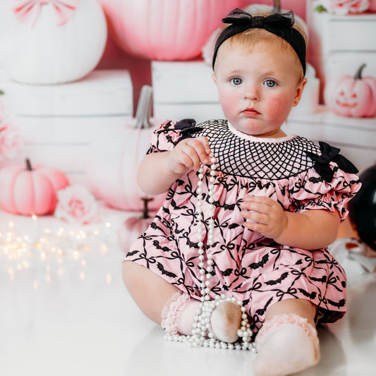 Toddler girl wearing pink hand smocked bats bloomer set with black bows by festive pastel Halloween pumpkins.