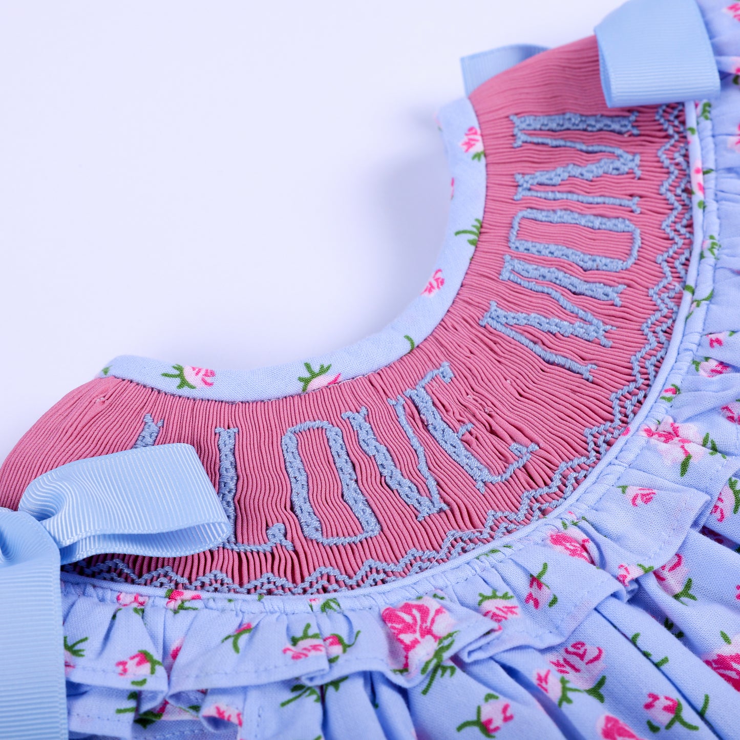 Detailed view of pink hand-smocked neckline with blue I Love Mom embroidery on baby girl floral outfit.