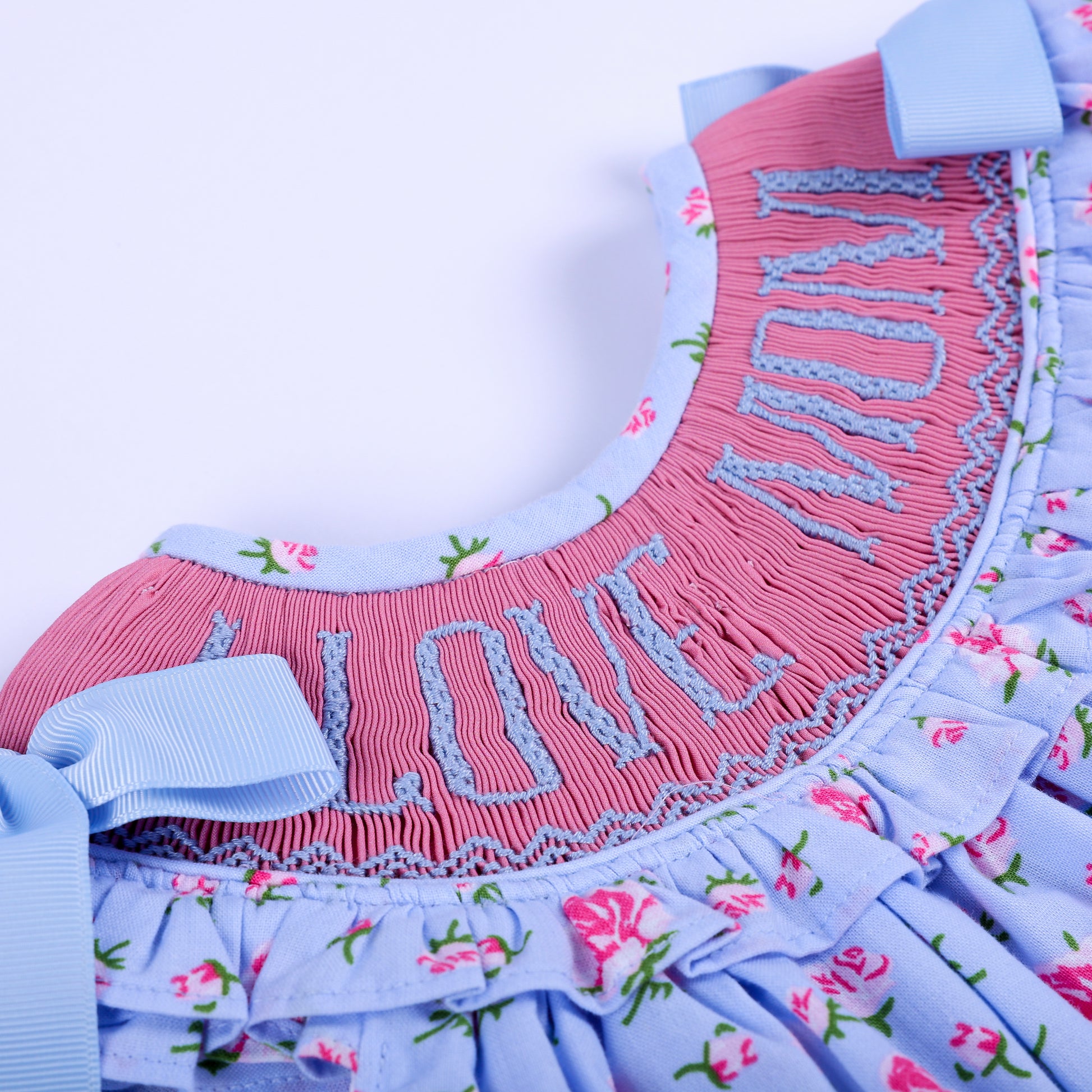 Detailed view of pink hand-smocked neckline with blue I Love Mom embroidery on baby girl floral outfit.
