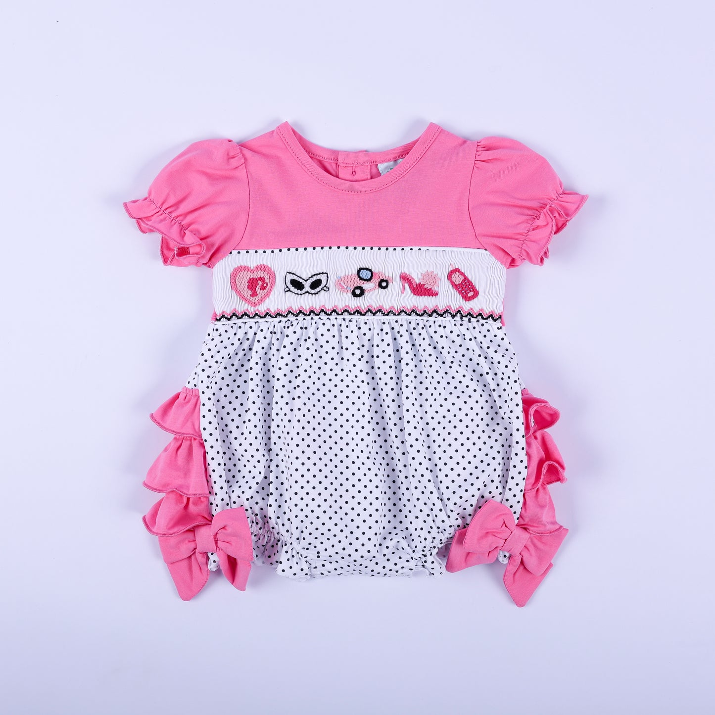 Pink polka dot hand-smocked baby girl bubble outfit featuring hand-embroidered fashion icons and ruffled sleeves.