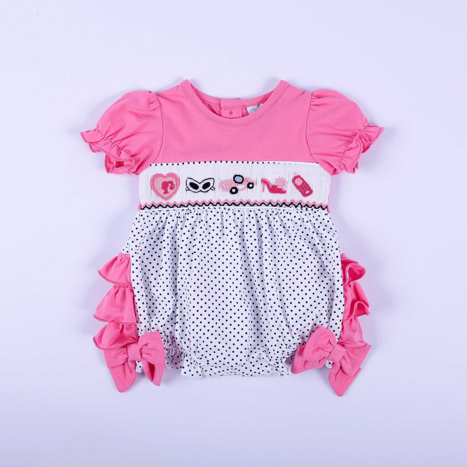 Pink polka dot hand-smocked baby girl bubble outfit featuring hand-embroidered fashion icons and ruffled sleeves.