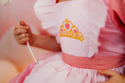 Detail of a smocked pink princess dress bodice with gold crown embroidery and ruffle trim, child holding a wand.