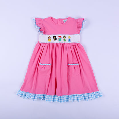 Full front view of girls pink princess knit hand smocked dress featuring blue gingham ruffle trim and pockets.