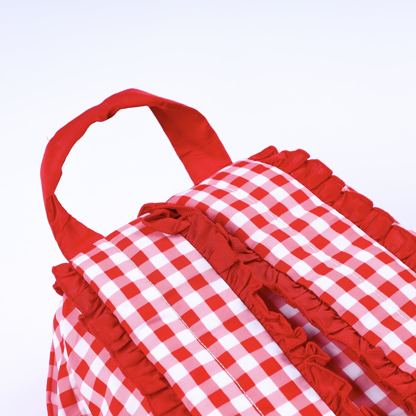 Close up top view of kids red gingham backpack featuring sturdy red carrying handle and ruffled straps.