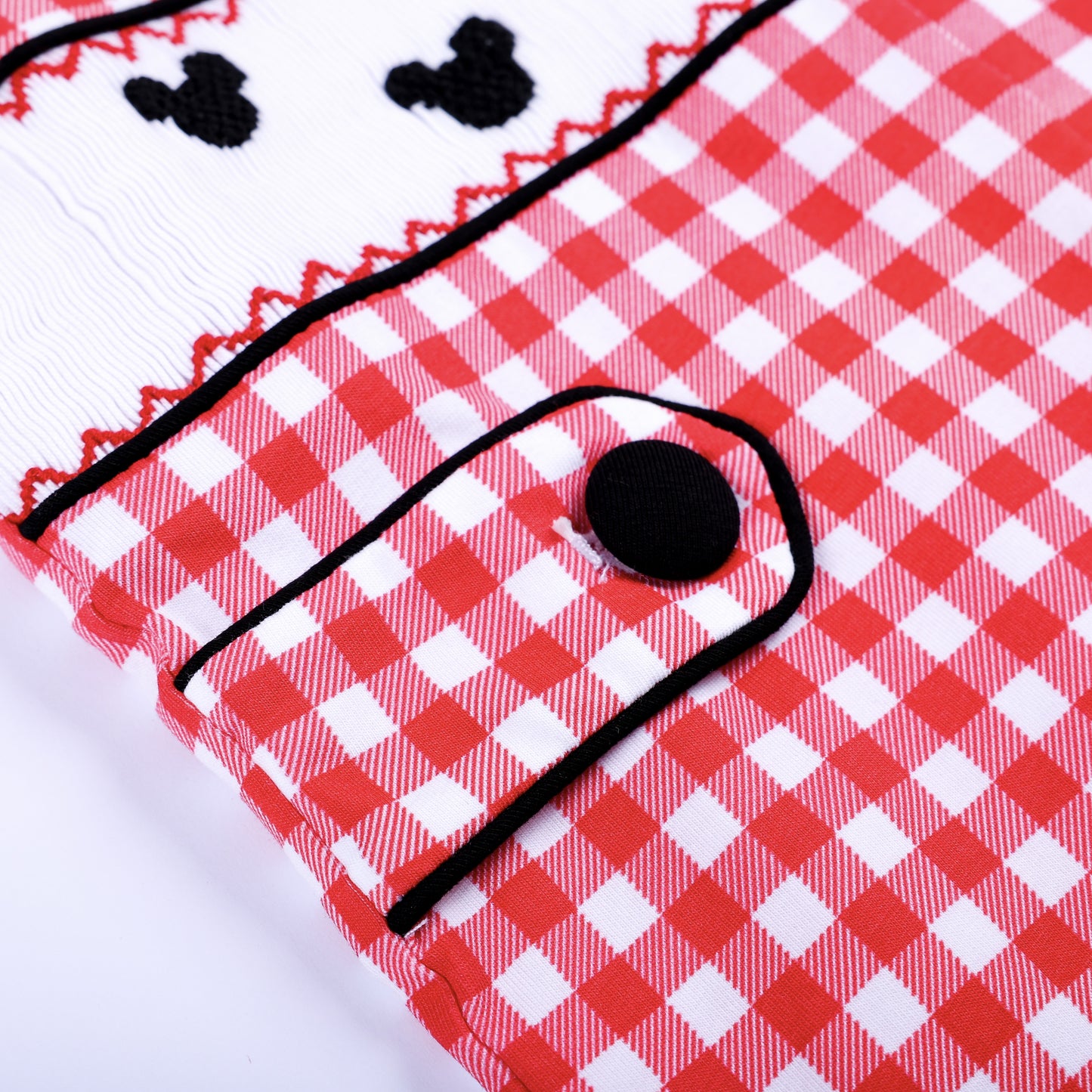 Macro detail of faux pocket flap featuring black edge trim and button on a red gingham smocked boys jon jon romper.