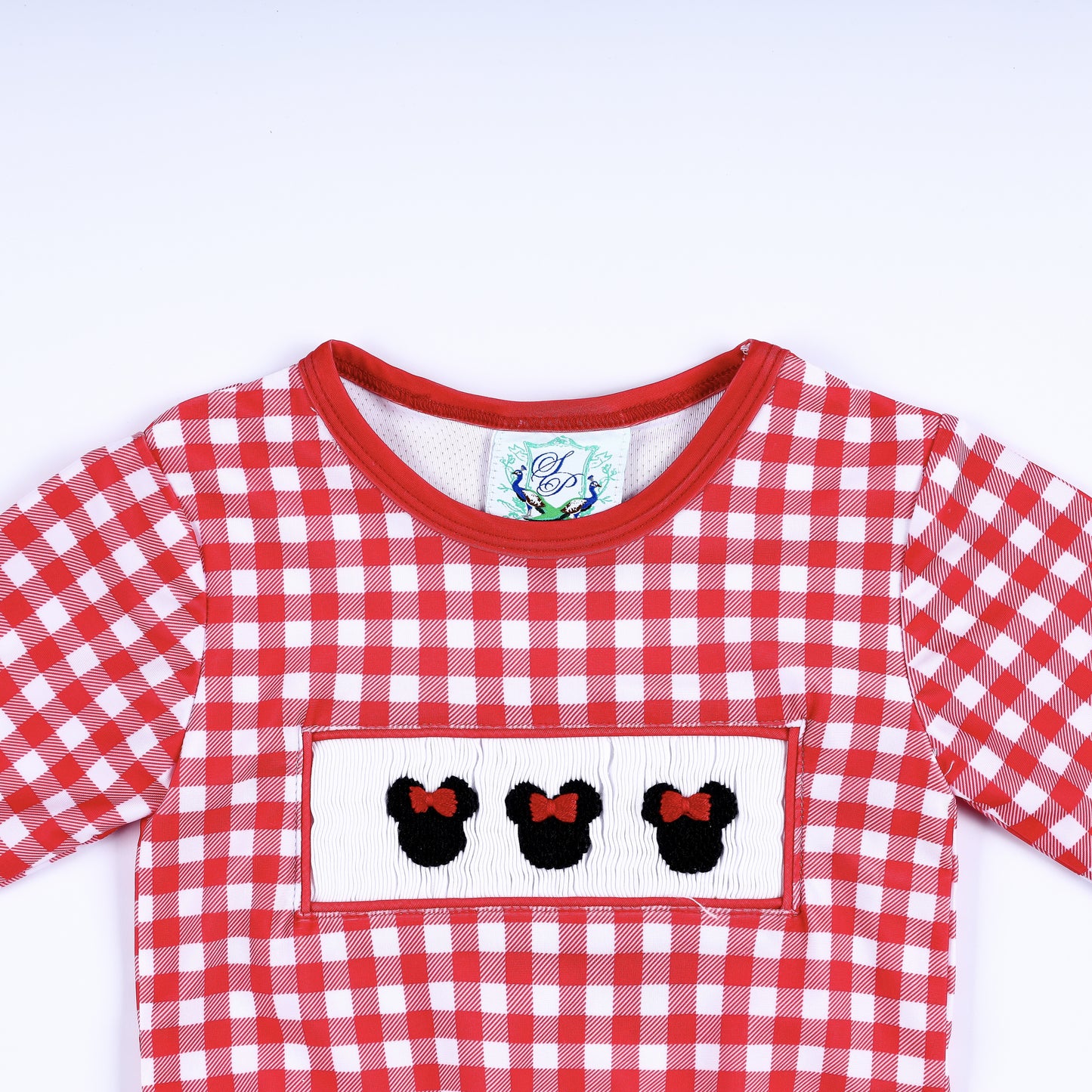 Close-up top view of red gingham long sleeve rash guard shirt with hand smocked Minnie Mouse bow embroidery plate.