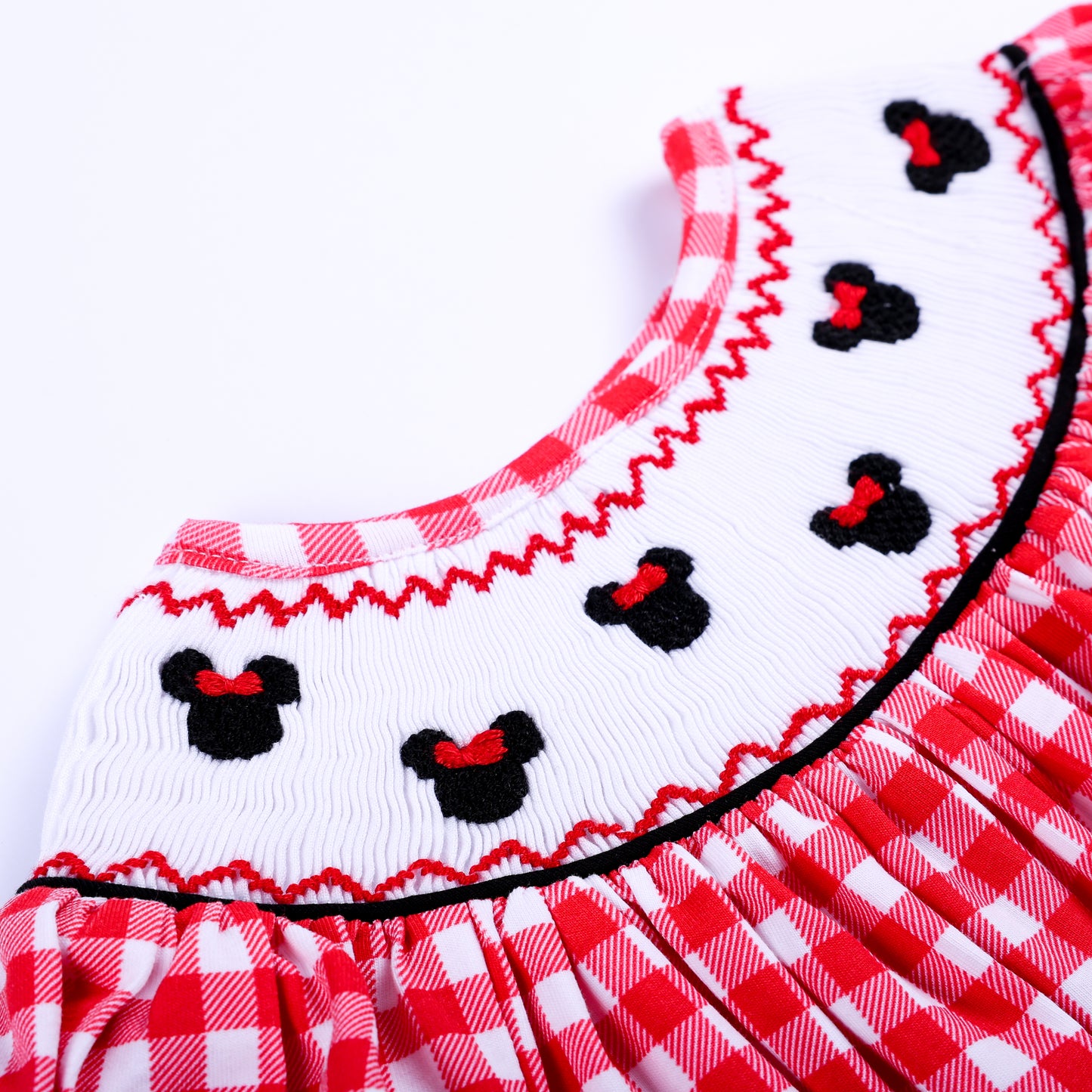 Detailed zoom of white hand smocked neck plate featuring black embroidered mouse icons with red bows on red gingham.