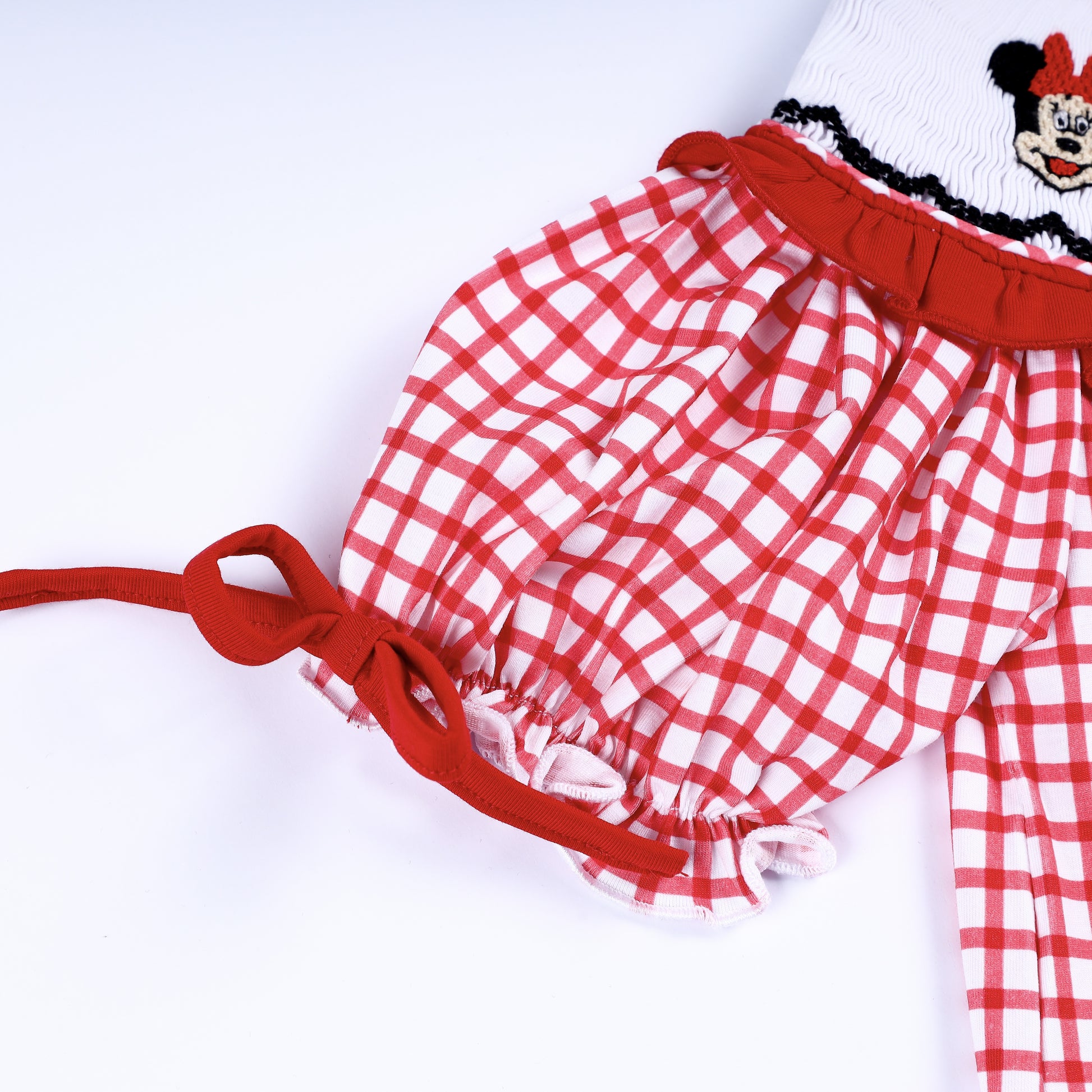 Close up of short puff sleeve on red gingham smocked dress featuring adjustable ribbon tie and ruffled trim.