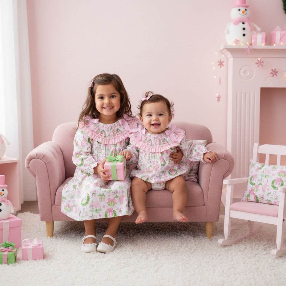 Two sisters on a pink couch wearing matching girls watercolor smocked Christmas dress and baby bubble outfit.
