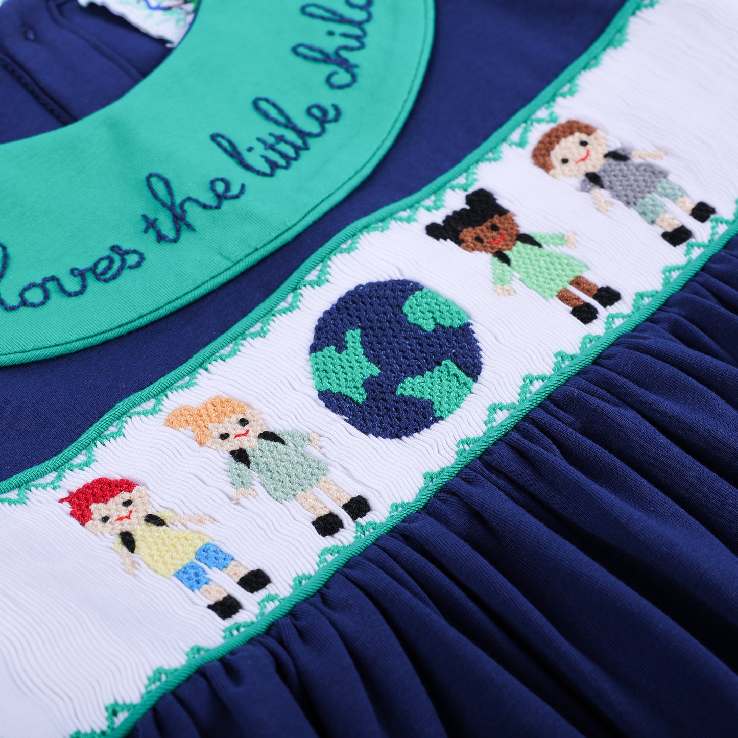 Close-up of the navy 'Jesus Loves the Little Children' dress bodice, showing the hand-smocked globe and diverse children.