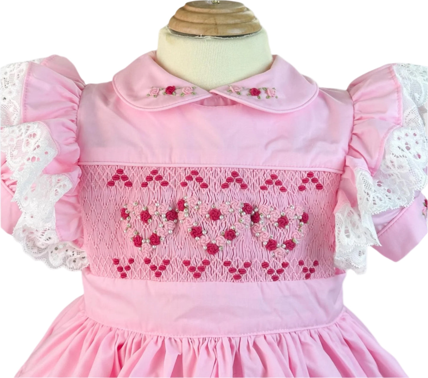 Close-up of the Vintage Valentine Heirloom Dress bodice by Smocked Peacock, showcasing intricate hand-smocked heart patterns with pearl details, an embroidered rosebud collar, and lace flutter sleeves.
