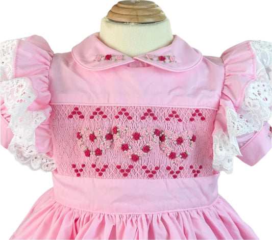 Close-up of the Vintage Valentine Heirloom Dress bodice by Smocked Peacock, showcasing intricate hand-smocked heart patterns with pearl details, an embroidered rosebud collar, and lace flutter sleeves.