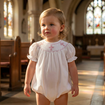 Baby girl in a white smocked bubble romper with pink hand-embroidered crosses on the neckline, standing in a church aisle.