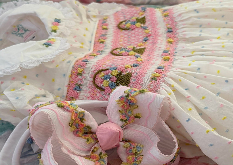 A close-up of a white embroidered dress with a pink smocked bodice, multicolor floral embroidery.