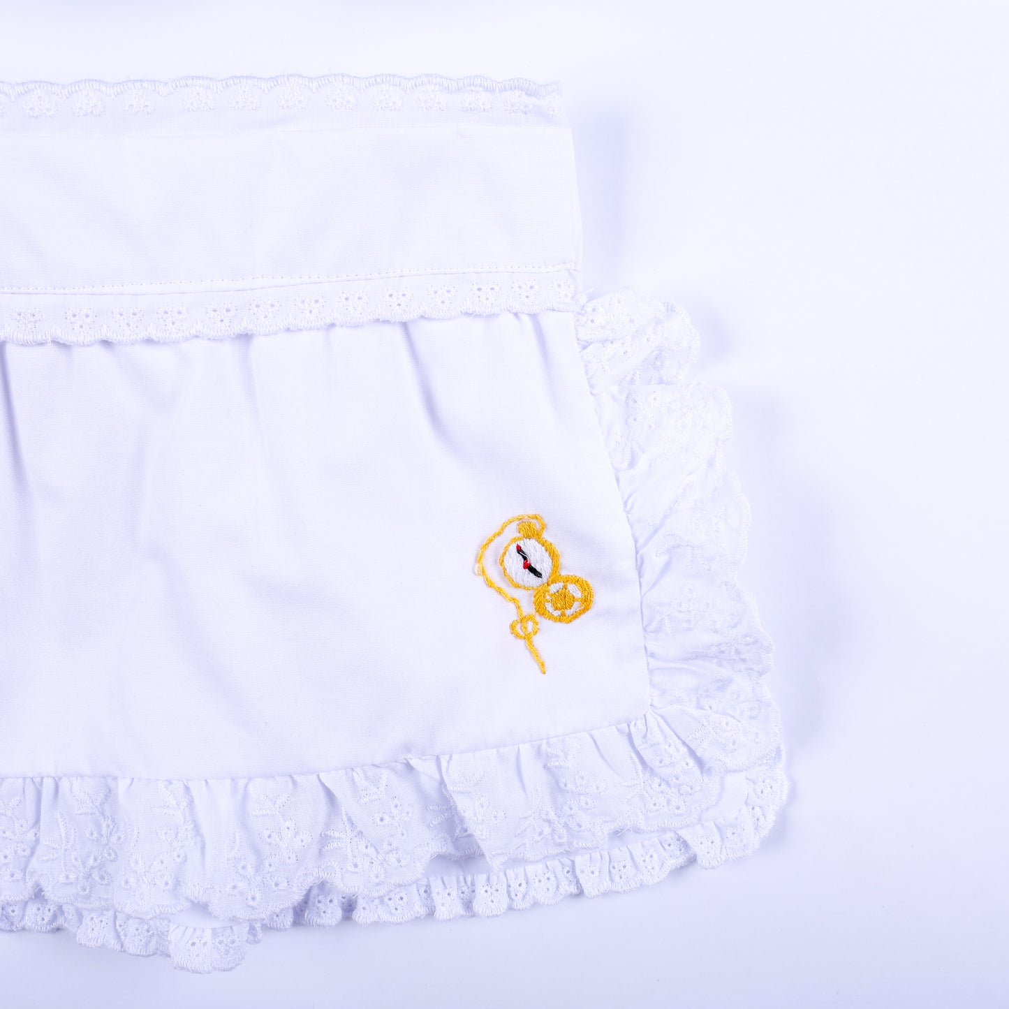 Detailed view of white eyelet lace ruffled apron featuring hand embroidered golden pocket watch motif.
