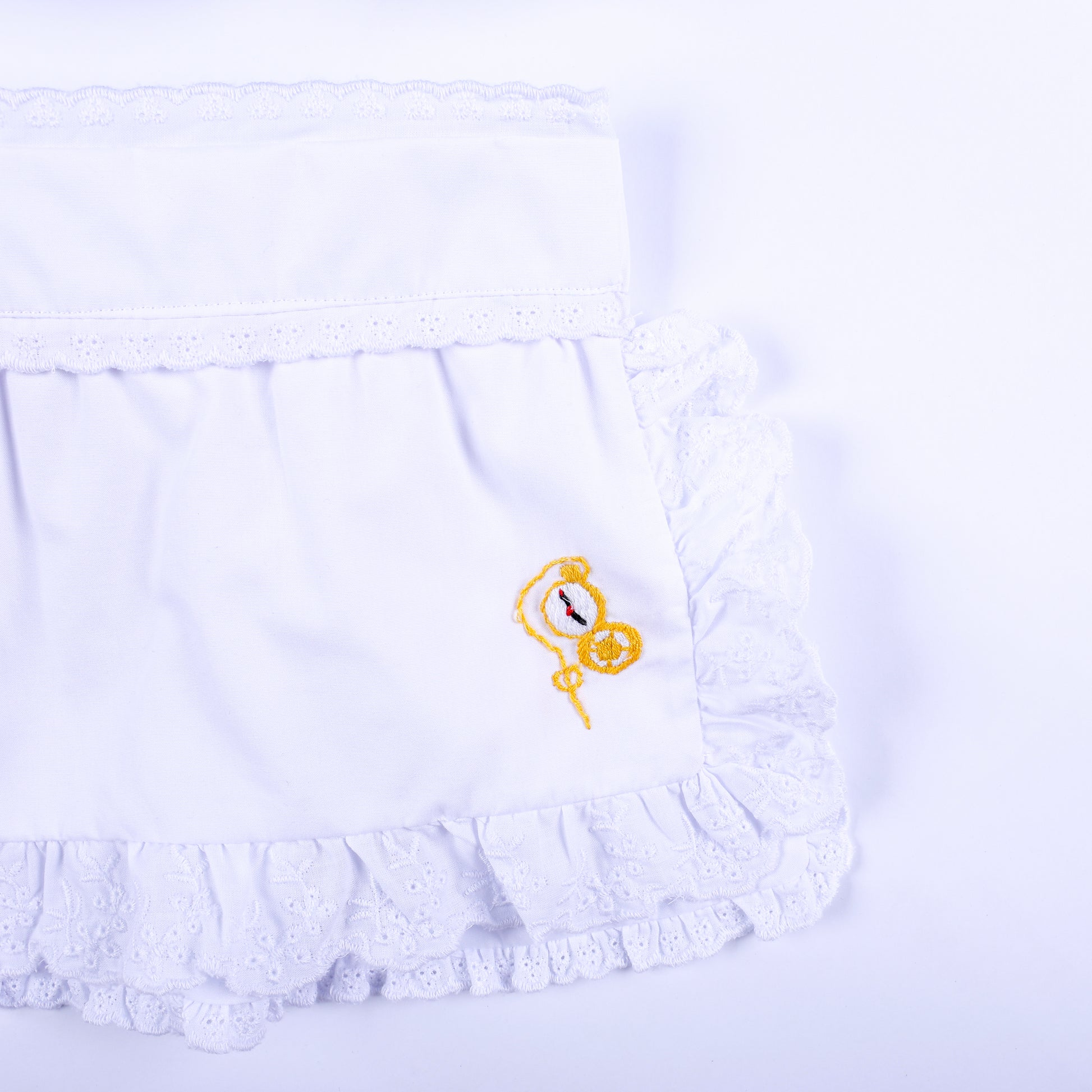 Detailed view of white eyelet lace ruffled apron featuring hand embroidered golden pocket watch motif.