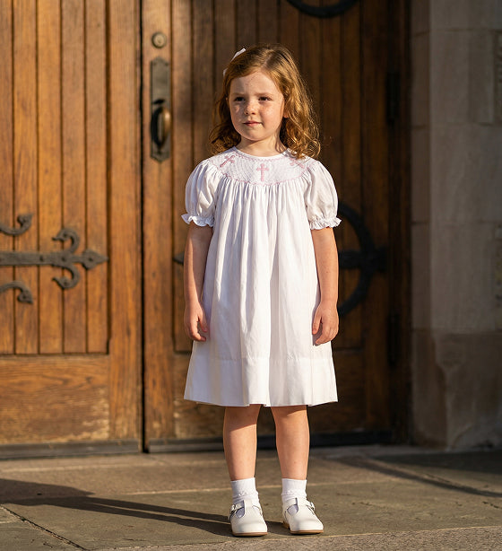 Little girl wearing a Sunday Best white smocked dress with embroidered pink crosses in front of church doors.