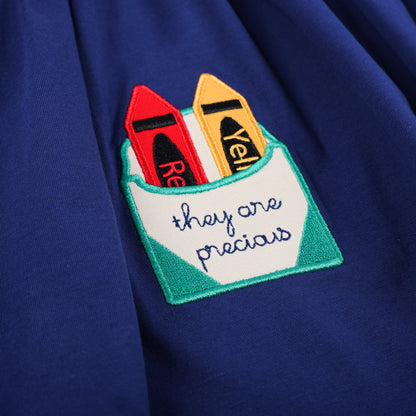 Detail of the crayon box pocket on the navy smocked dress, embroidered with 'they are precious' and holding crayons.