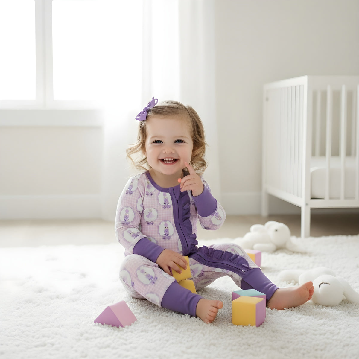 Smiling toddler sitting on playroom carpet wearing the purple Nutcracker ballet ruffle girls zippy pajamas.