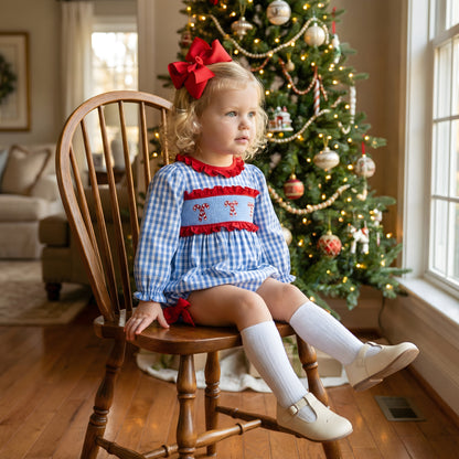 Toddler girl wearing a blue gingham hand-smocked candy cane bubble by a Christmas tree.