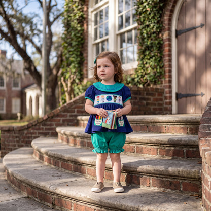 Toddler girl wearing a blue smocked top with an embroidered collar and matching teal bloomers.
