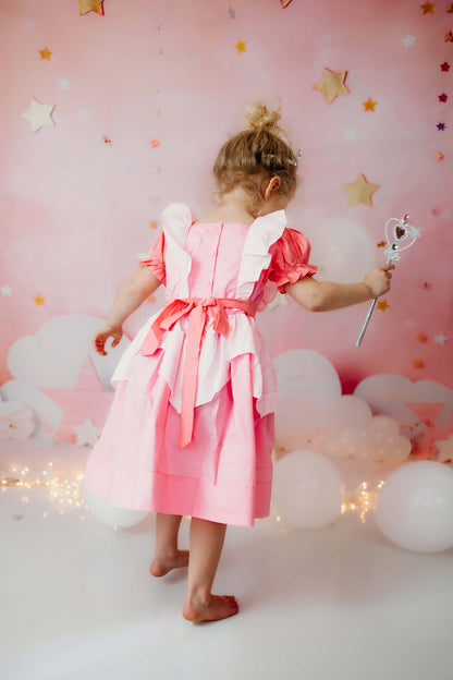 Back view of a barefoot toddler girl in a pink princess dress with a tied sash bow and puff sleeves, holding a scepter.