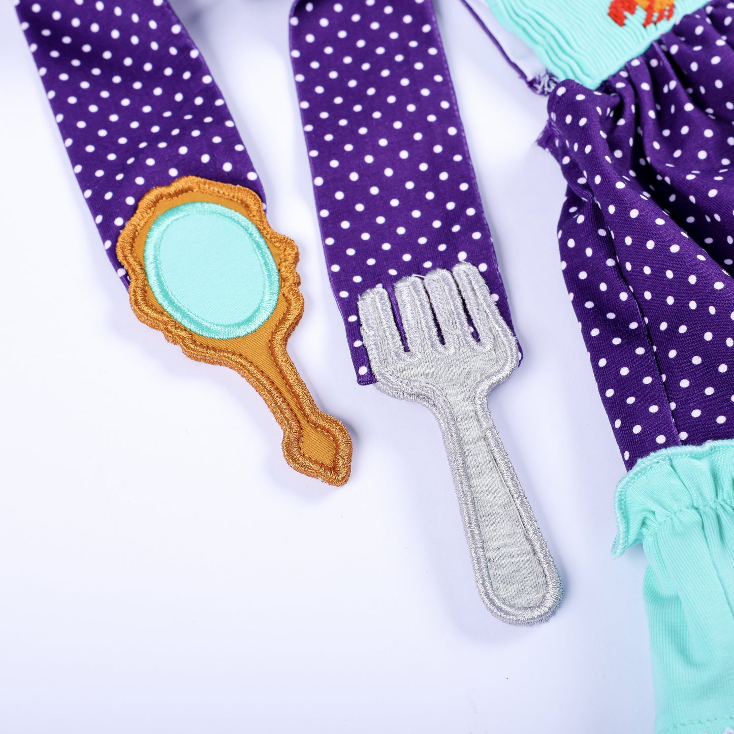 Detailed view of hand embroidered mirror and fork appliques on the shoulder straps of smocked bloomer set.