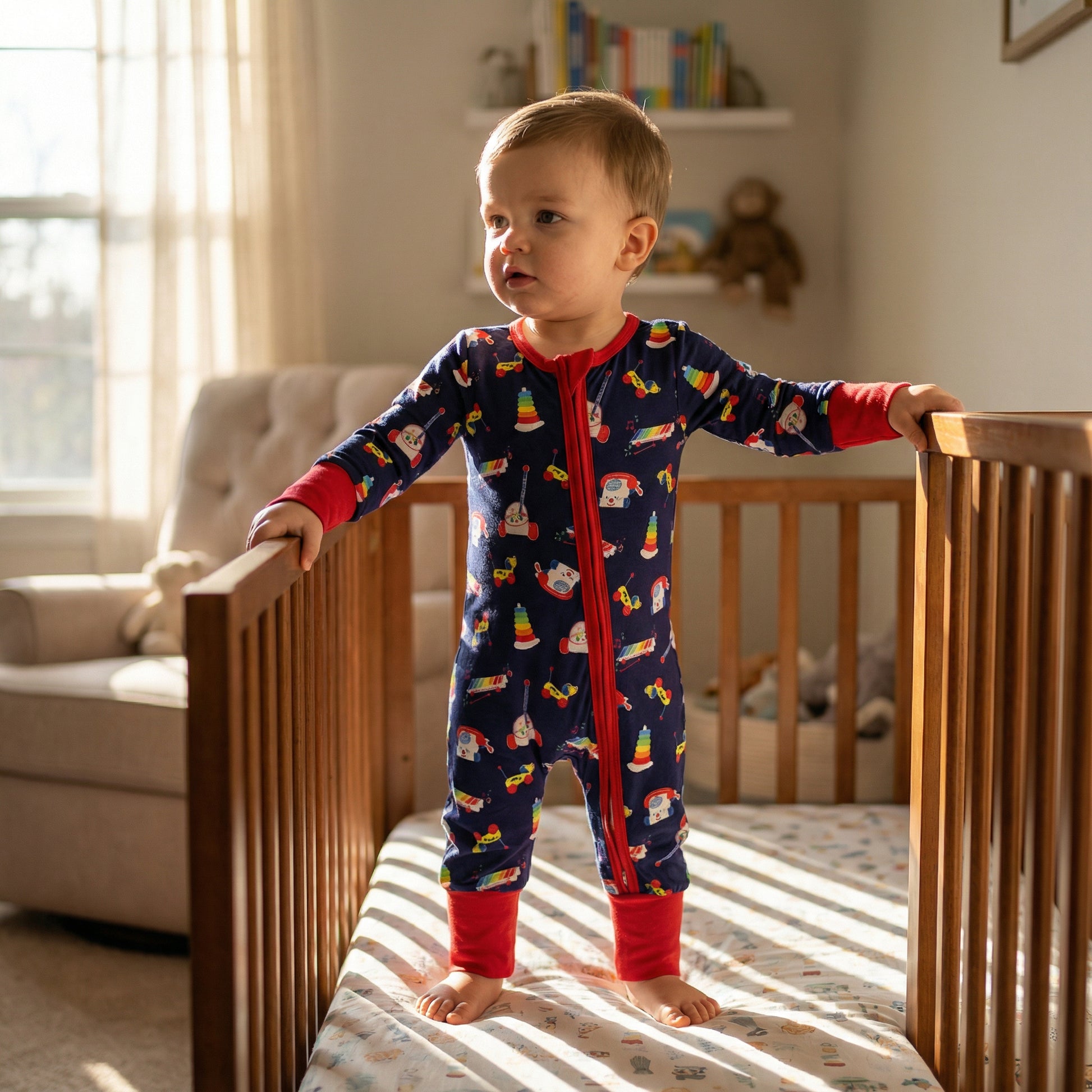 Toddler boy standing in crib wearing navy zippy pajamas with vintage 80s and 90s toy print.