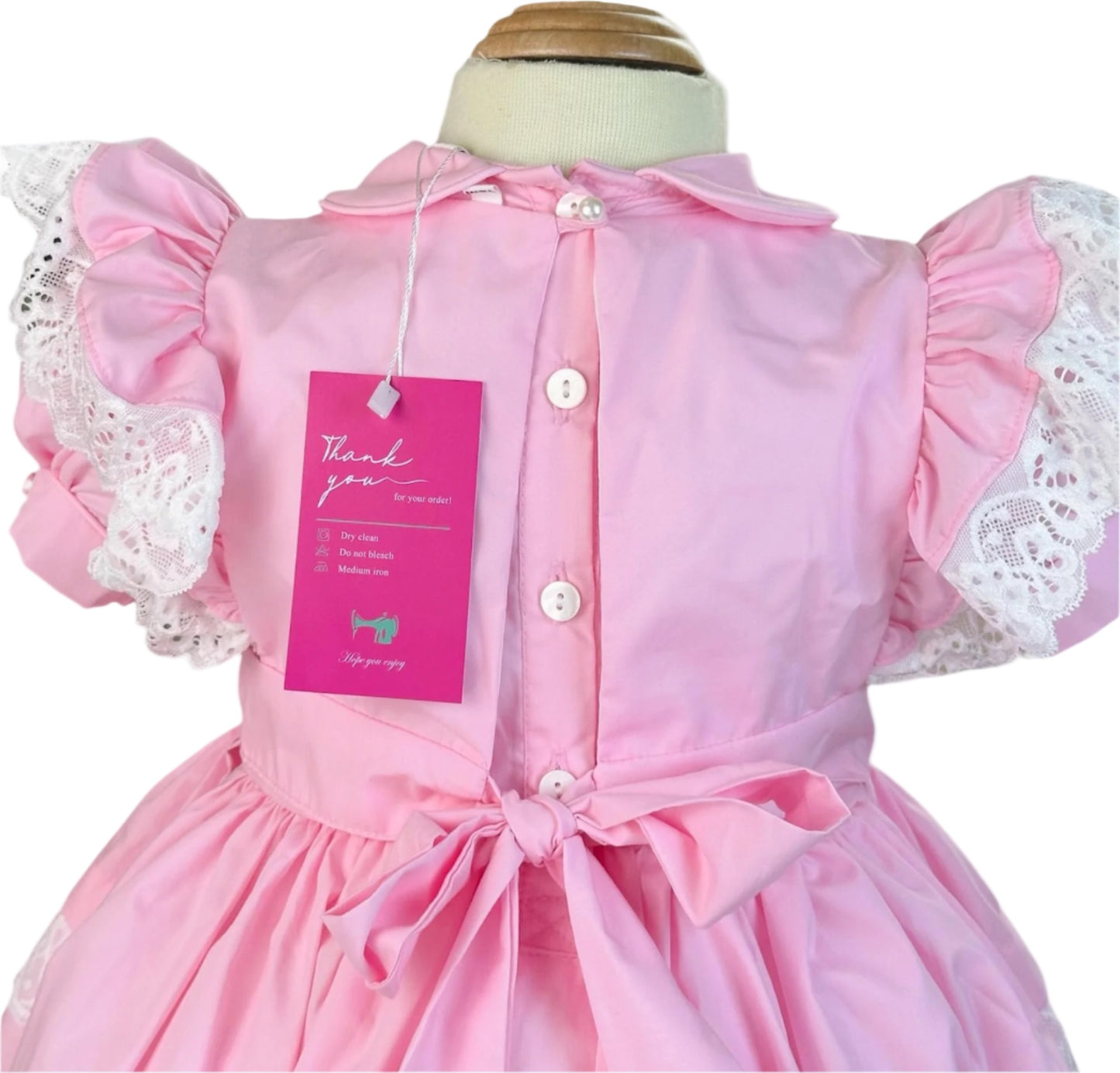 Back view of the pink Vintage Valentine Heirloom Dress by Smocked Peacock, featuring a classic white button-down closure, a large tied waist bow, and lace-trimmed flutter sleeves.